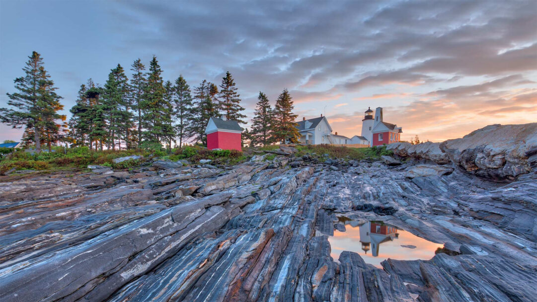 A captivating 4K wallpaper showcasing Pemaquid Point Light on the Maine coast, situated above a rugged rocky shore during sunset. The dramatic sunset sky paints the scene with warm hues, reflected in a foreground tide pool that mirrors the lighthouse and illuminates the ancient, layered rocky formations.