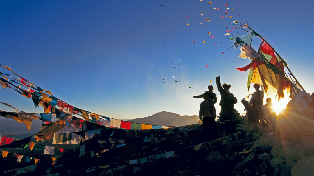 A powerful 4K wallpaper presenting a group of pilgrims silhouetted against a mountain sunset, actively throwing colorful wind horses into the clear blue sky. Vibrant prayer flags ripple across the rocky foreground, their colors intensified by the golden sunburst on the horizon, evoking a sense of spiritual celebration and vastness.