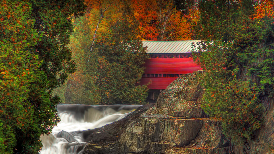 A picturesque 4K wallpaper showcases the iconic Pont Rouge Red Bridge gracefully spanning above the powerful Palmer River waterfall during autumn. Its striking red structure beautifully complements the dynamic rush of the waterfall and the rich spectrum of vibrant orange, yellow, and deep green autumn trees surrounding the riverbanks.