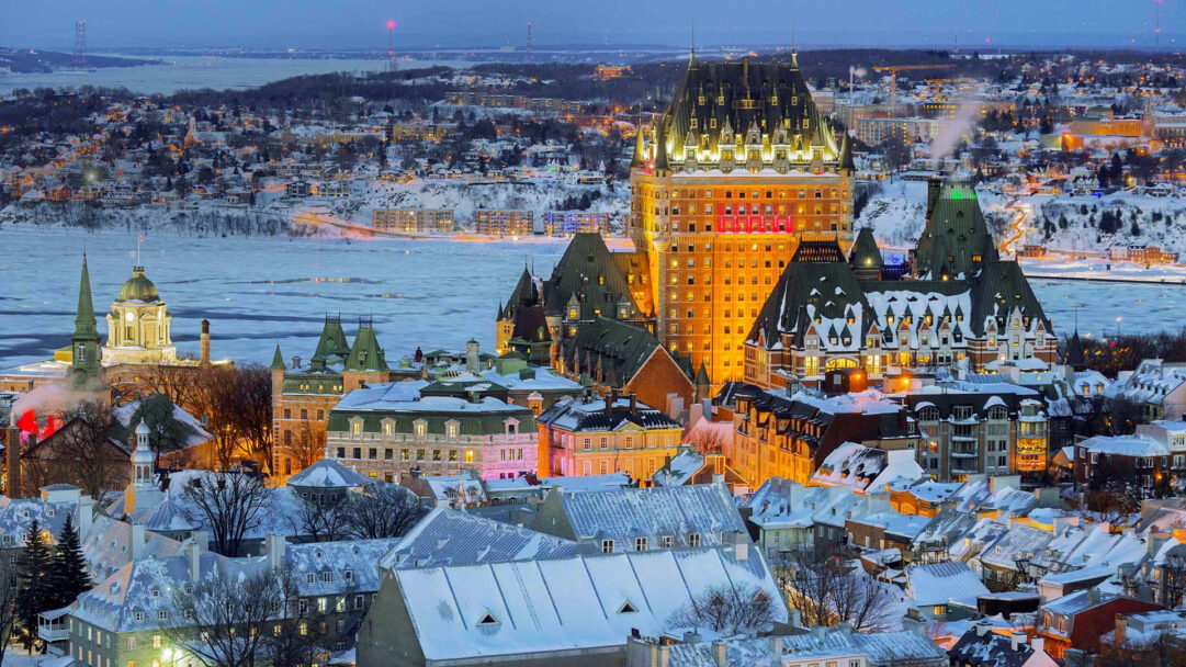 A picturesque 4K wallpaper capturing the Quebec City Old Town skyline during a vibrant winter night. The iconic Château Frontenac glows with warm golden light, contrasting beautifully with the cool blue twilight, snow-dusted roofs, and frozen St. Lawrence River, evoking a serene yet vibrant winter mood.