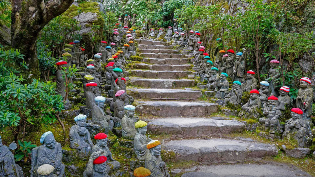 A delightful 4K wallpaper showcases numerous Rakan statues lining a moss-covered stone staircase at the Daisho-in Temple on Miyajima Island, Japan. The cheerful array of multi-colored knitted hats adorning each statue offers a striking visual element, bringing a playful warmth to the ancient, verdant temple environment.