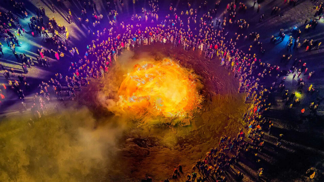 An immersive 4K wallpaper portraying a vibrant New Year's Eve bonfire celebration in Reykjavik, 2014, with a vast crowd gathered in a wide circle. The immense, roaring bonfire emits a powerful golden-orange light, brilliantly illuminating the surrounding attendees and filling the dark night with warmth and festive energy.