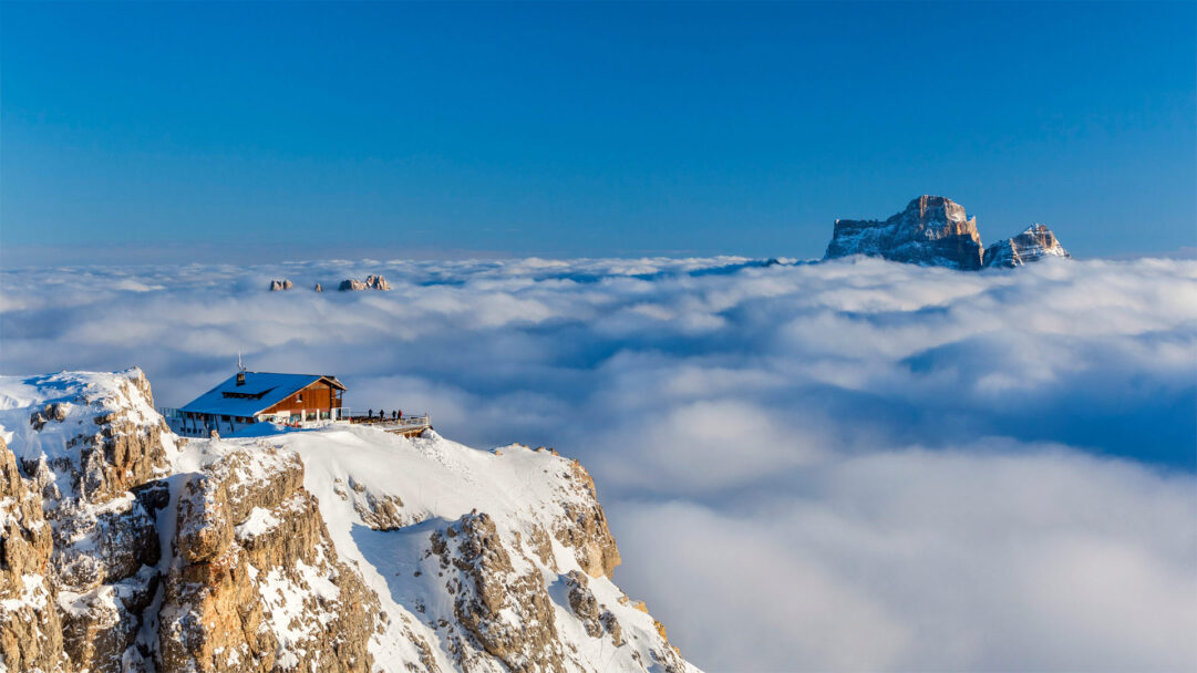 An expansive 4K wallpaper depicting Rifugio Lagazuoi, a cozy mountain hut perched on a snow-covered peak, rising above a tranquil sea of winter clouds. In the distance, the rugged, snow-dusted peak of Monte Pelmo emerges majestically from the cloudscape, bathed in golden sunlight under a brilliant blue sky.