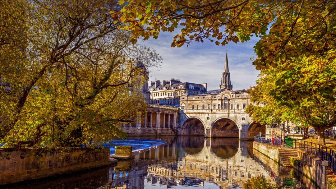 A picturesque 4K wallpaper showcasing the iconic Pulteney Bridge gracefully spanning the River Avon in Bath, England, during a vibrant autumn. Golden autumn leaves frame the historical architecture, reflecting beautifully in the calm river waters under a bright sky, evoking a serene and timeless atmosphere.