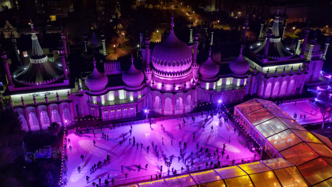 A mesmerizing 4K wallpaper showcasing the Royal Pavilion Ice Rink in Brighton, England, bustling with skaters under the night sky. The iconic Royal Pavilion glows with vibrant purple illumination, reflecting dazzling light onto the ice and creating a festive, energetic mood.
