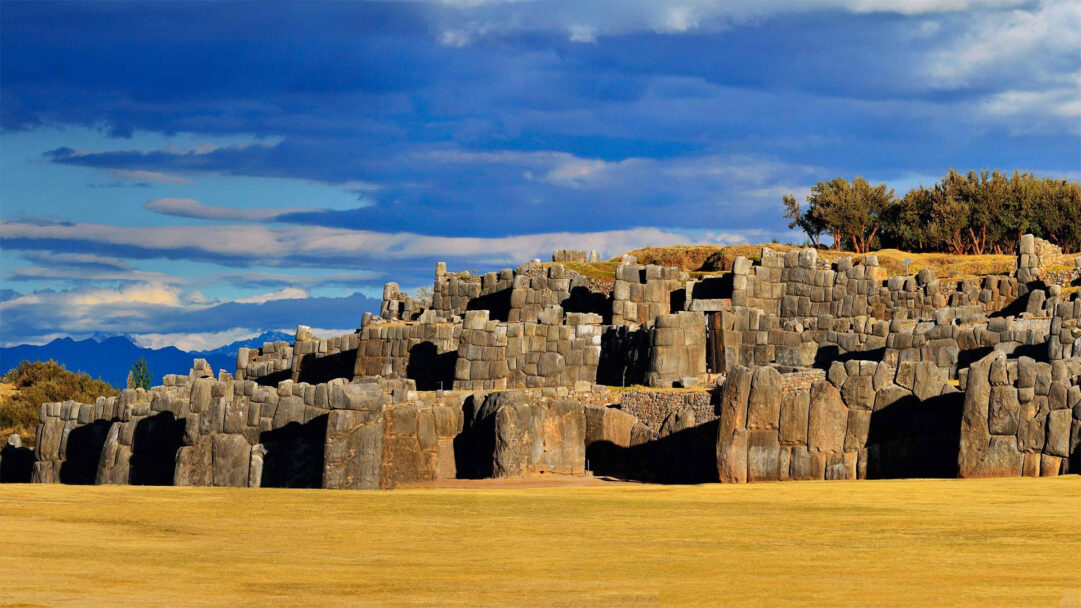 An awe-inspiring 4K wallpaper showcasing the monumental Sacsayhuaman Inca Fortress in the rugged landscape of Cusco, Peru. Massive, intricately fitted stone walls rise from a sun-drenched golden field under a dramatic blue sky with sweeping clouds, conveying ancient power and grandeur.