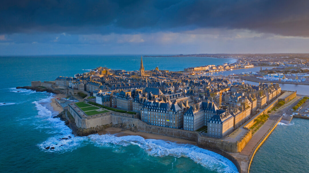 An impressive 4K wallpaper captures an aerial view of the historic Saint-Malo Walled City on the Brittany, France coast, surrounded by the deep blue ocean. Golden light illuminates sections of the ancient fortifications and buildings, creating a vivid contrast against the crashing waves and the dramatic, cloud-filled sky above.