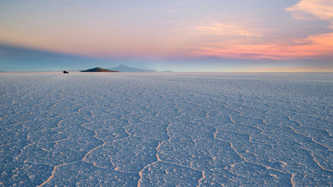 A stunning 4K wallpaper featuring the immense Salar de Uyuni salt flat in Bolivia at sunset, with a distant dark island under a vast sky. Warm golden light illuminates the intricate polygonal patterns of the salt crust, contrasting with the soft pastel pinks and blues of the expansive sky.