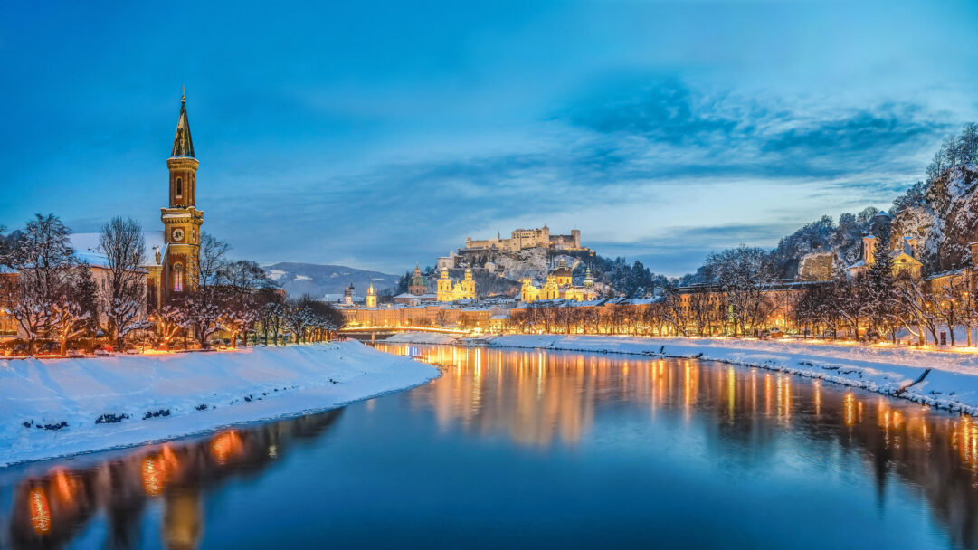 A captivating 4K wallpaper of Salzburg, Austria, showcasing a beautiful winter cityscape with the Salzach River winding through it and the majestic Hohensalzburg Fortress perched above. The warm golden lights of the snow-dusted city cast enchanting reflections upon the deep blue river, creating a serene and picturesque twilight scene.