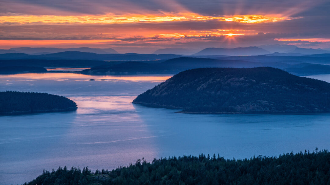 An evocative 4K wallpaper captures the San Juan Islands, Washington, as a sprawling landscape of tree-covered islands and tranquil waters. The dramatic sunset bathes the horizon in brilliant orange and gold, with visible sun rays piercing through clouds and casting a serene, reflective glow across the deep blue waterways.