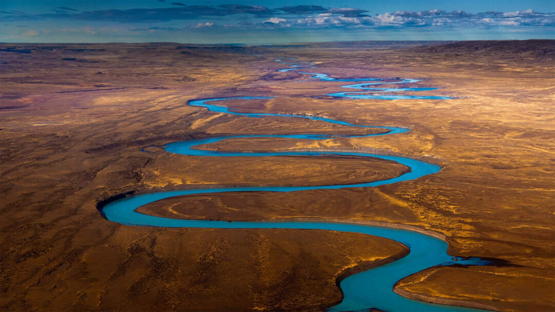 An expansive 4K wallpaper revealing the Santa Cruz River winding through the vast, arid Patagonian landscape of Argentina. Its vibrant turquoise waters cut a dramatic, serpentine path across the golden-brown plains, creating a stunning visual contrast under a partly cloudy sky.