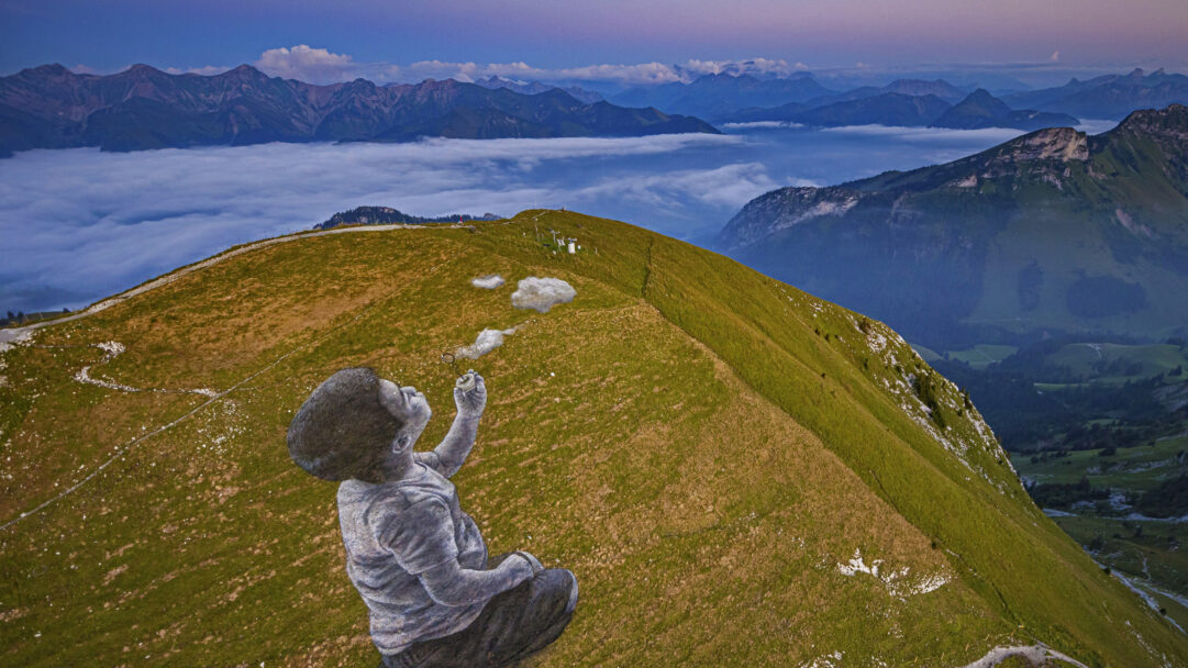 A monumental 4K wallpaper showcases Saype's 'A New Breath' land art, featuring a vast painting of a child blowing bubbles across the verdant Moléson-sur-Gruyères Mountain slope. The child looks up as the white 'bubbles' merge with the real clouds filling the valley below, creating an awe-inspiring scene against the serene backdrop of distant peaks under a lavender twilight sky.