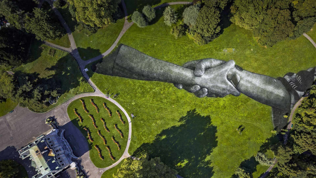 An expansive 4K wallpaper presents Saype's 'Beyond Walls' Handshake Art Installation stretching across the verdant grounds of Parc de la Grange. Its immense, hyperrealistic stone-like hands interlock on the natural canvas, symbolizing unity and connection within the bright park environment.