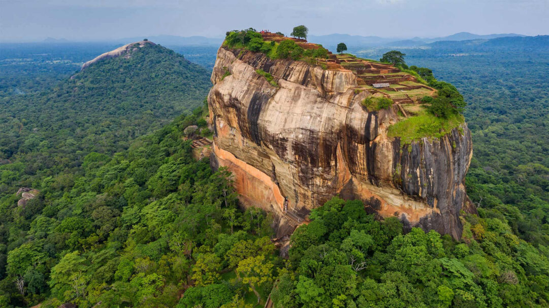 An awe-inspiring 4K wallpaper showcasing the colossal Sigiriya Rock Fortress, rising majestically from a dense, verdant jungle in Sri Lanka's Central Province. Its sheer, multi-hued cliffs bear ancient structures and contrast dramatically with the sprawling green canopy, hinting at a timeless, powerful presence.