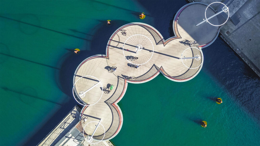 A captivating 4K wallpaper presents an aerial view of Copenhagen, Denmark’s unique Circle Bridge, where cyclists traverse its series of interlocking circular platforms over brilliant turquoise water. Strong sunlight creates striking elongated shadows of the bridge's masts and active cyclists, emphasizing the modern design and vibrant urban scene.