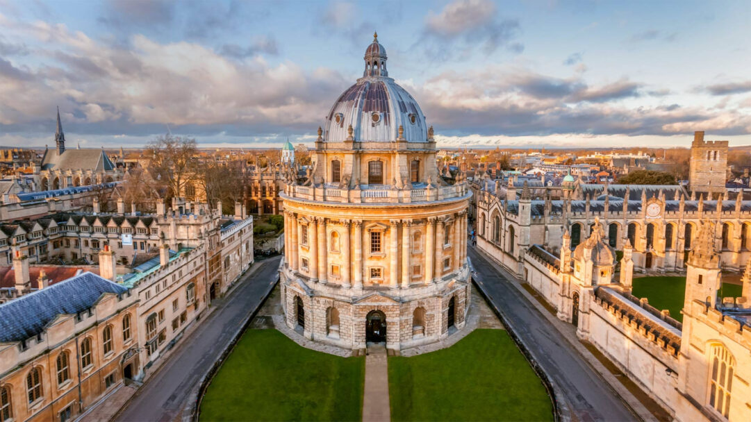 A majestic 4K wallpaper featuring The Radcliffe Camera, a grand domed building at the heart of historic Oxford, England, surrounded by intricate collegiate architecture. Golden light bathes the detailed stone facades, highlighting its classical grandeur against a dramatic, cloudy sky.