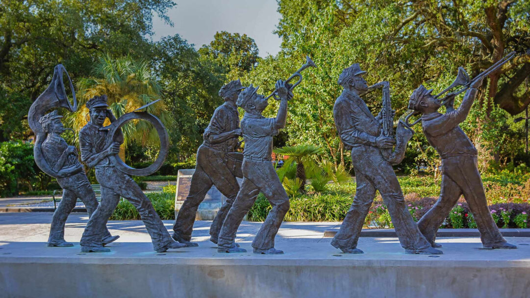 A vibrant 4K wallpaper showcasing The Roots of Music Cultural Sculpture Garden in Louis Armstrong Park, New Orleans, where a procession of bronze musicians marches, playing various instruments. The intricate bronze figures, depicting lively musicians with a tuba, trumpet, saxophone, and trombone, are bathed in warm sunlight against a backdrop of verdant trees and subtropical foliage, evoking the rich musical heritage of the city.