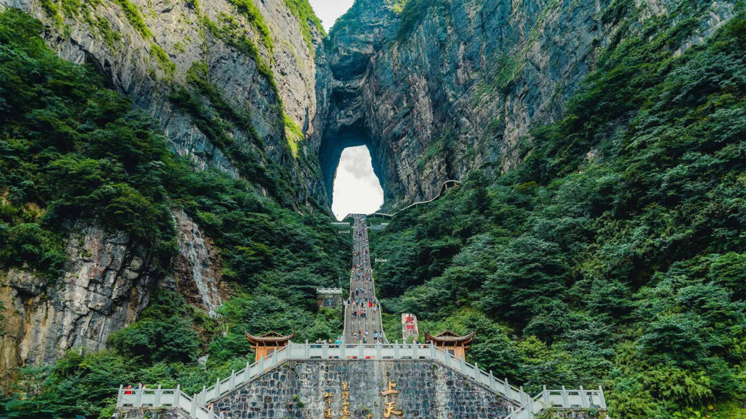An awe-inspiring 4K wallpaper captures the famous Heaven's Gate at Tianmen Mountain in Zhangjiajie, China, showcasing a grand stairway carved into towering, verdant cliffs. The seemingly endless 'Stairway to Heaven' ascends dramatically through a narrow mountain pass to the iconic natural archway, bathed in bright light, emphasizing the monumental journey.