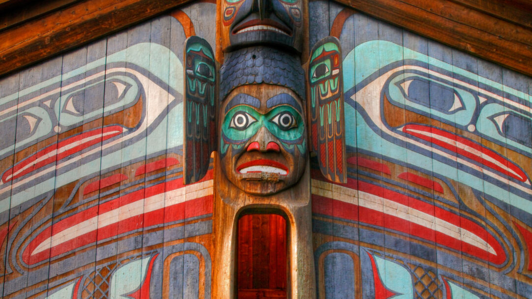 A captivating 4K wallpaper showcases a vibrant close-up of the Totem Bight Clan House Totem Pole Art in Ketchikan, Alaska. The pole's central carved face, featuring intricate teal, red, and black details against the natural wood grain, emanates a powerful cultural presence.