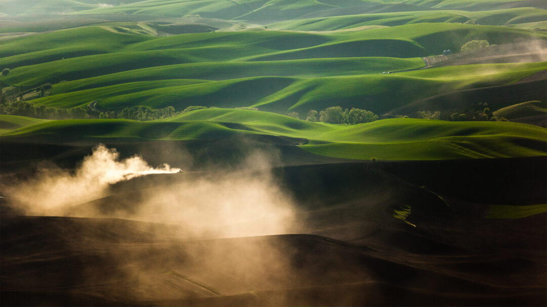 A captivating 4K wallpaper presents a tractor tilling the undulating Palouse fields near Pullman, Washington. Golden sunlight pierces through vast plumes of dust rising from the freshly tilled soil, creating a striking contrast with the verdant, rolling hills stretching into the distance.
