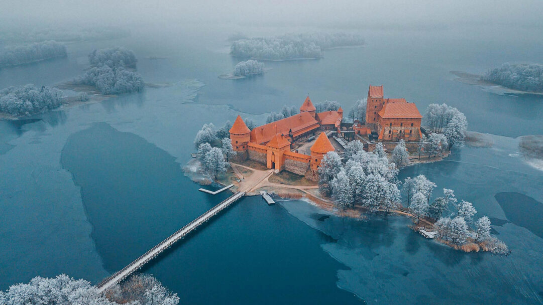 A captivating 4K wallpaper presents Trakai Island Castle Museum in a serene winter aerial view, nestled on an island amidst a partially frozen lake. Its distinctive orange-red brick structures stand in striking contrast to the frosty white trees and the cool, icy blue water, all veiled in a soft winter mist.