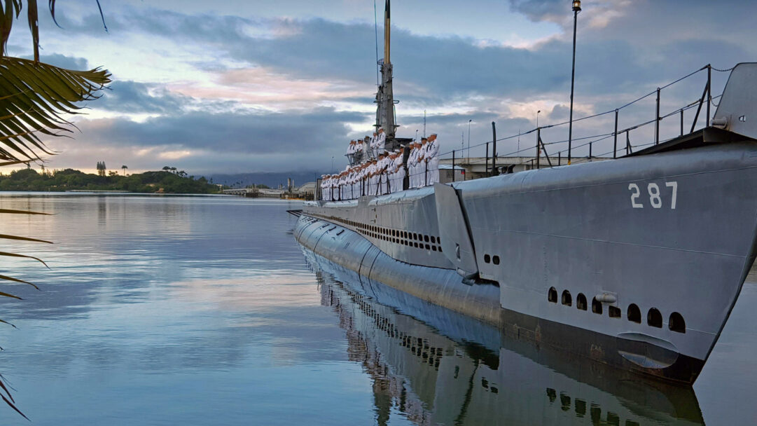 An evocative 4K wallpaper capturing US Navy sailors standing at attention on the deck of the USS Bowfin, a WWII Museum Submarine, docked in tranquil harbor waters. The calm surface beautifully reflects the vessel and the soft, cloud-streaked sky, enhancing the solemn and majestic atmosphere of this historic scene.