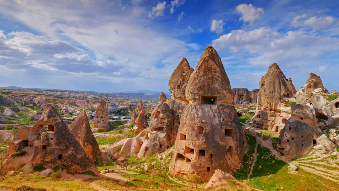 An enchanting 4K wallpaper unveils the unique Uçhisar landscape, prominently featuring ancient fairy chimneys intricately carved with cave dwellings across rolling hills. The striking interplay of sunlight and shadow highlights the warm, earthy textures of the formations against vibrant green foliage and an expansive blue sky, evoking a sense of historical wonder.