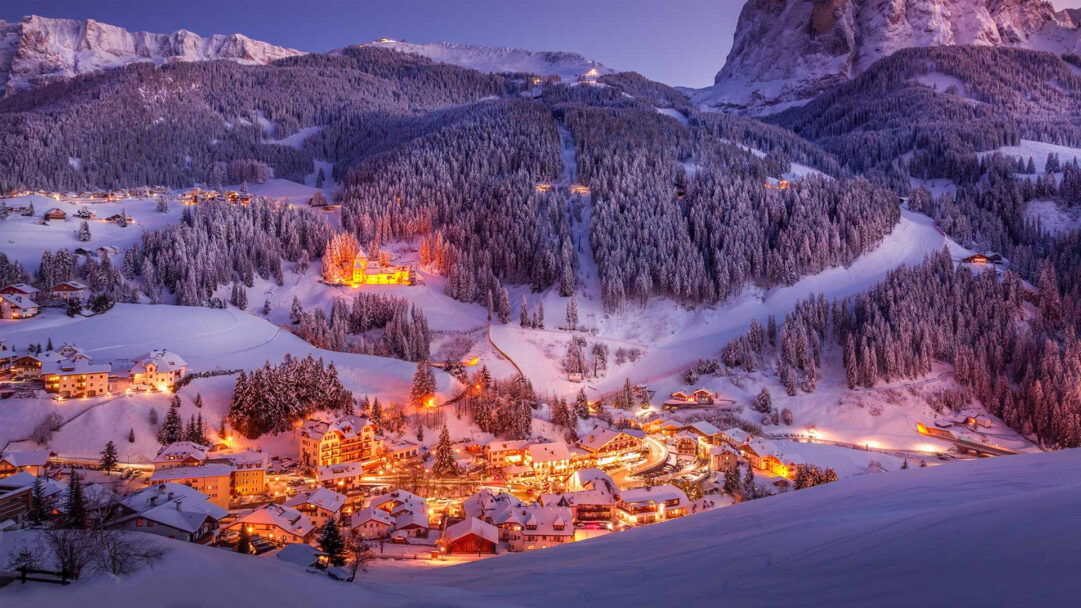 An enchanting 4K wallpaper depicts the Val Gardena winter village nestled within the snow-covered Dolomites at sunset. Warm, golden lights from the buildings glow invitingly against the deep blue twilight sky and pristine white snow, creating a serene, magical winter atmosphere.