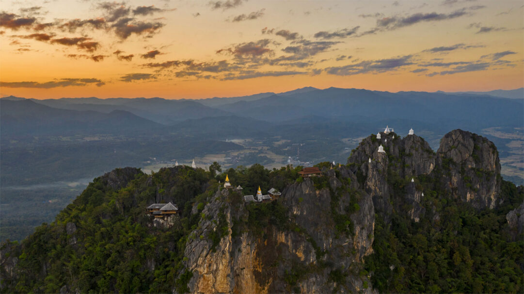 A majestic 4K wallpaper showcasing the Wat Chaloem Phra Kiat Phrachomklao Rachanusorn Temples perched dramatically on rugged mountain peaks. The scene is bathed in the warm, golden light of sunset, casting a serene glow over the distant mountains and highlighting the pristine white chedis against the fading sky.