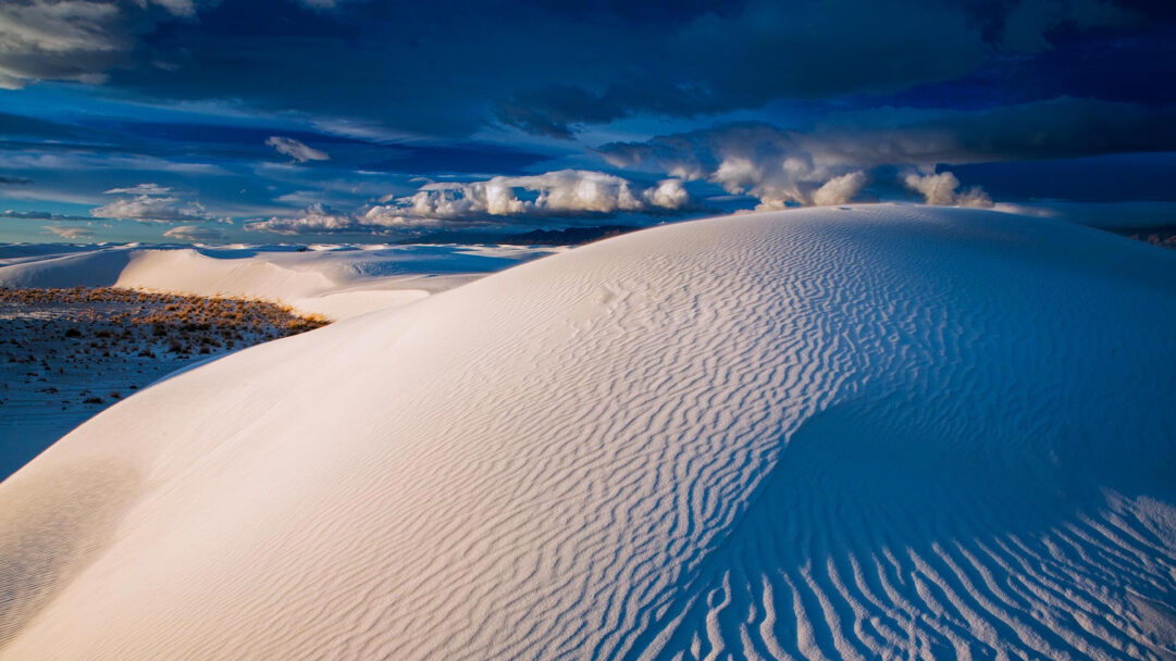 A captivating 4K wallpaper showcasing the expansive gypsum dunes of White Sands National Park under a dramatic, cloud-filled deep blue sky. The crisp white gypsum forms elegant, rippled contours that glow under dynamic sunlight, sharply contrasting with the deep blues of the towering clouds overhead.