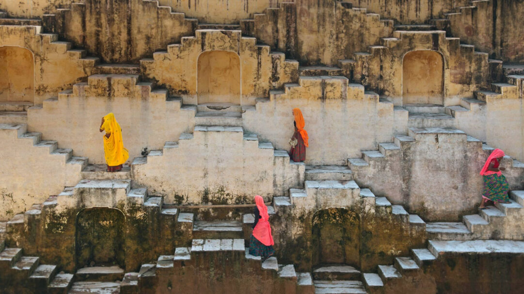 A captivating 4K wallpaper of women gracefully navigating the ancient, geometric steps of a stepwell at Amber Fort in Jaipur. Their vibrant traditional attire provides a striking contrast against the weathered, textured stone walls, creating a timeless and visually rich scene.