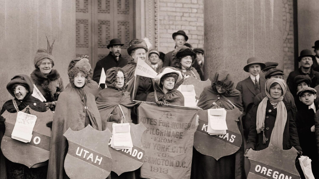 A momentous 4K wallpaper depicting a determined group of Women's Suffragists, including men and children, gathered with banners and state shields during their historic 1913 march from New York City to Washington D.C. The monochrome palette and the participants' earnest expressions, particularly the women cloaked against the elements, underscore their unwavering resolve and the profound historical significance of their Votes for Women pilgrimage.