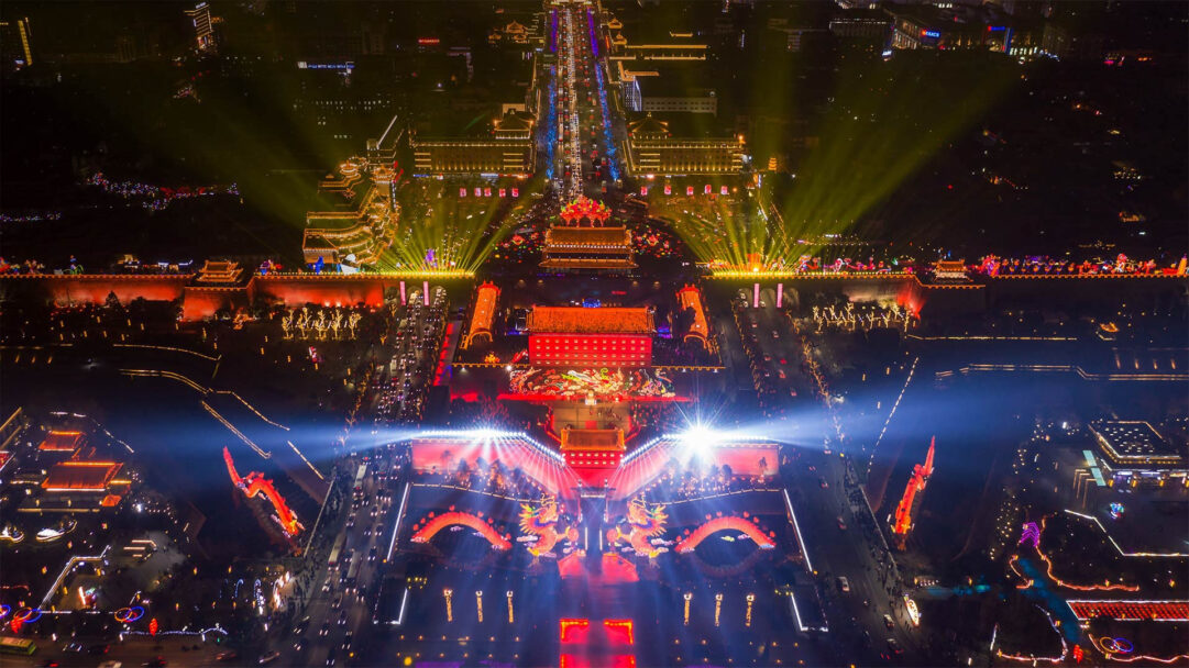 An electrifying 4K wallpaper presents the breathtaking aerial view of Xi'an's Chinese New Year celebration at night, showcasing grand traditional architecture and bustling streets. Vibrant red and golden lights illuminate intricate dragon sculptures and city gates, with powerful spotlights beaming into the dark sky, creating an atmosphere of immense joy and festivity.