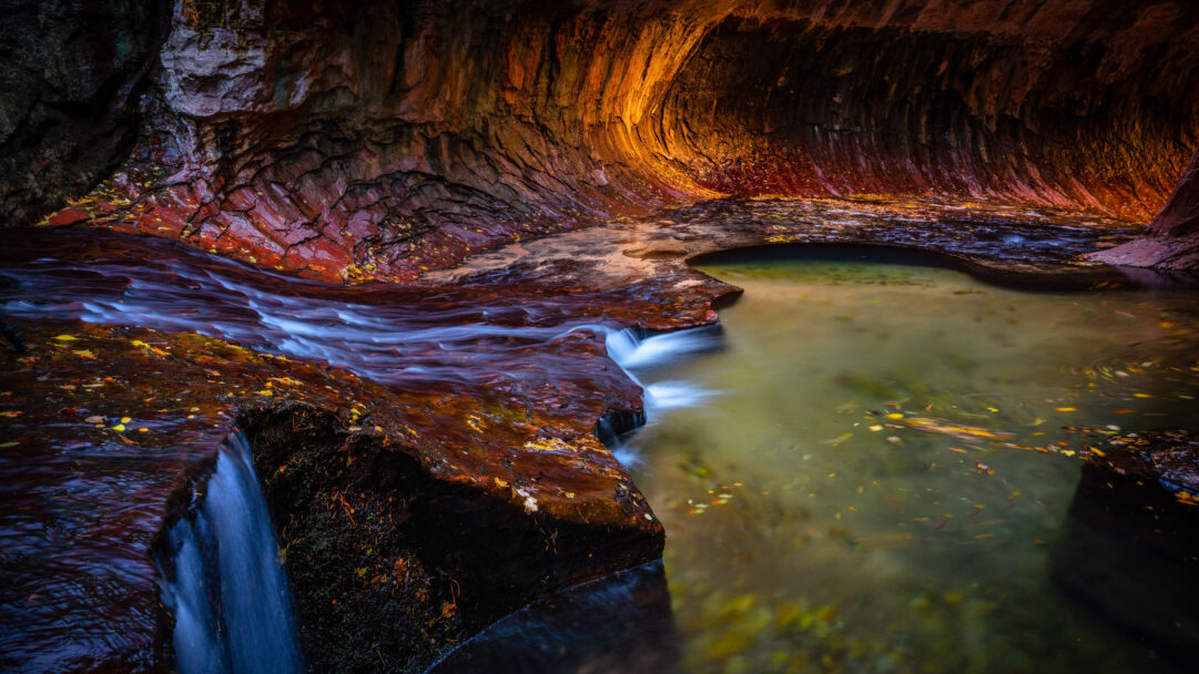 An immersive 4K wallpaper capturing a majestic waterfall stream flowing through The Subway Slot Canyon in Zion National Park. Ethereal long-exposure water cascades over smooth, reddish-brown rock formations, reflecting the warm, glowing light illuminating the sculpted 'Subway' walls and deep green pool.