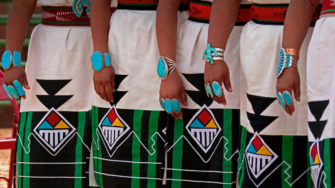 A captivating 4K wallpaper showcases Zuni Olla Maidens standing together in their exquisite Inter-Tribal Ceremonial traditional dress. The vibrant turquoise jewelry on their hands and wrists strikingly complements the black, green, and multi-colored geometric designs on their white skirts.